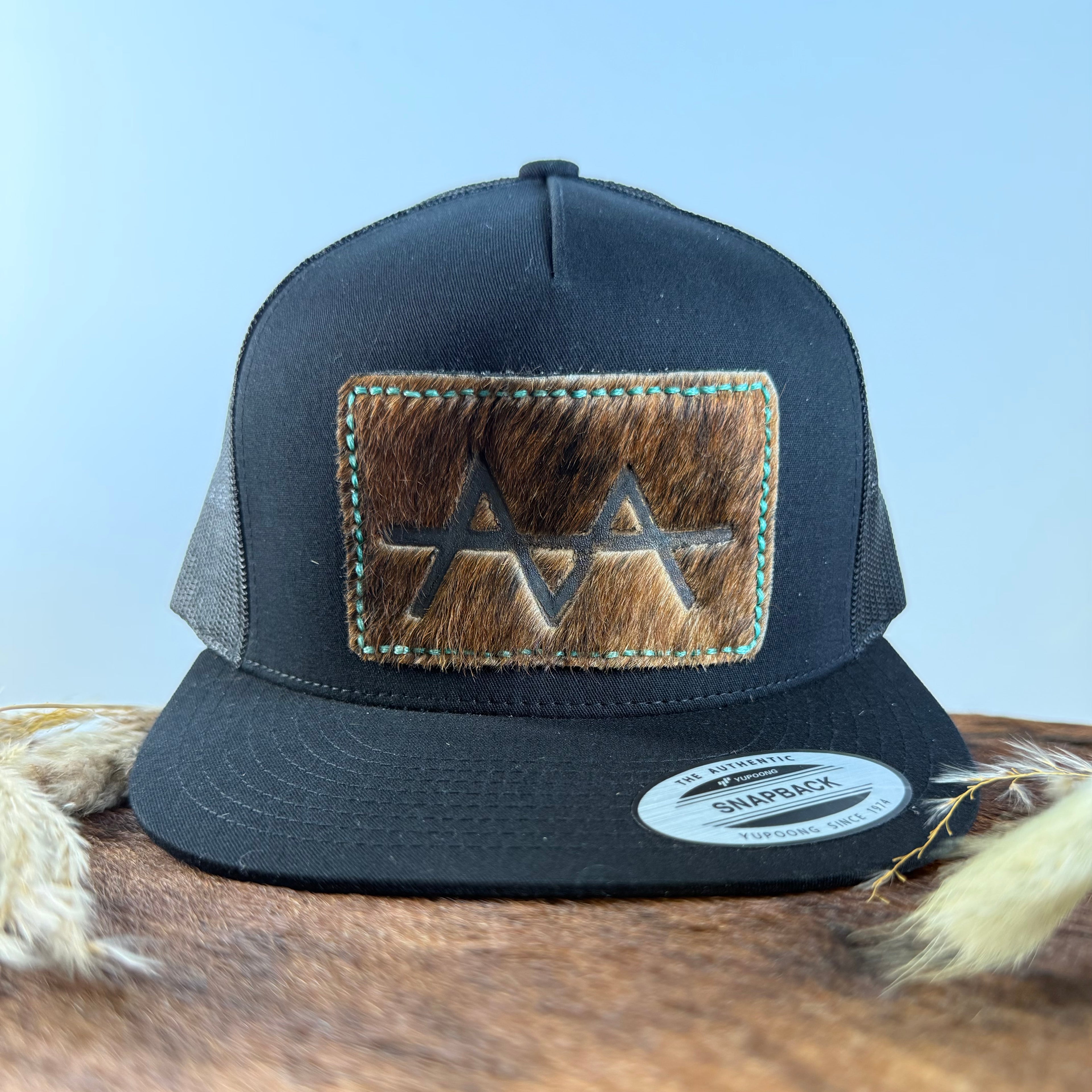 All-black trucker snapback hat with medium warm brindle cowhide patch and turquoise hand-stitched border, branded with a registered cattle brand