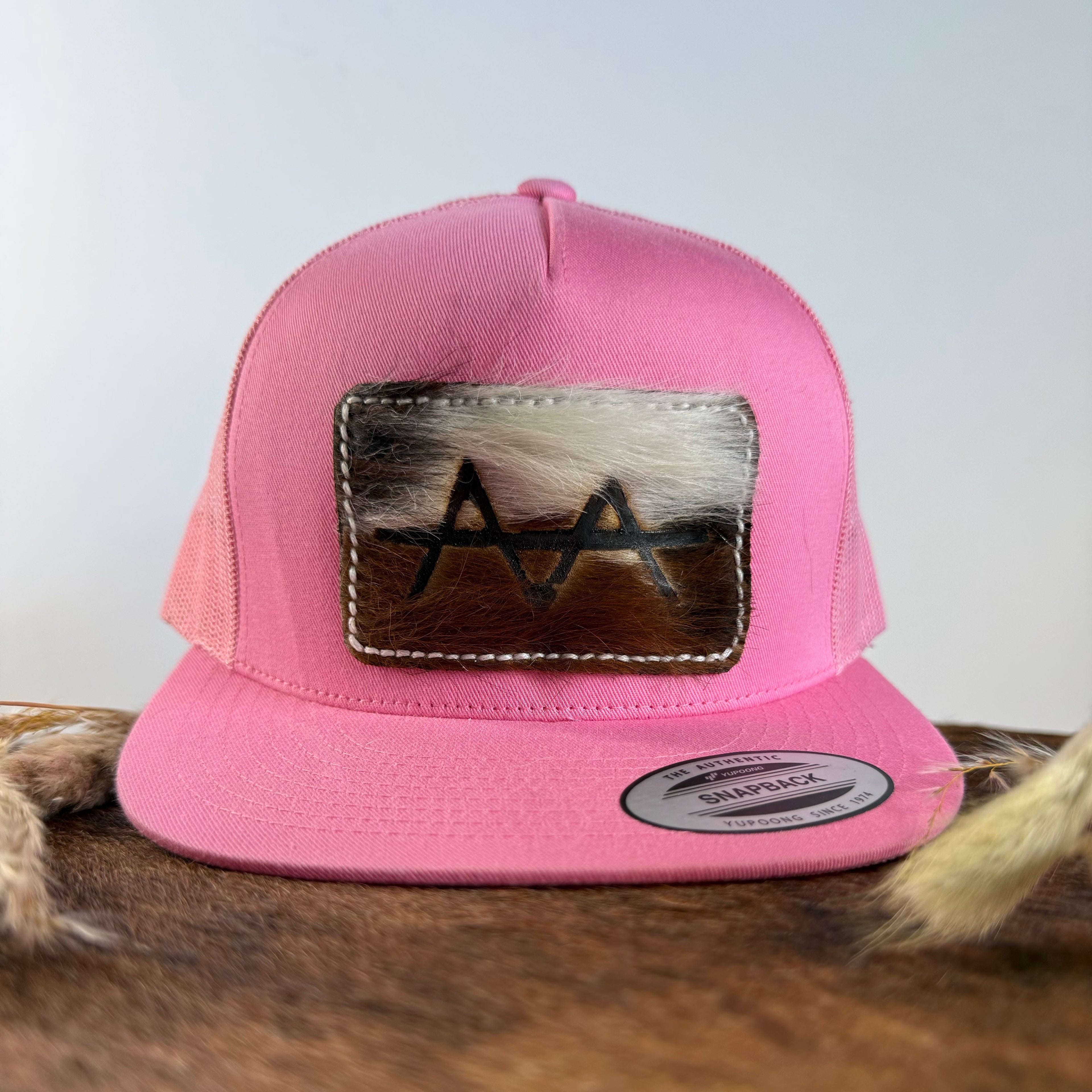 Pink trucker snapback hat with two-tone white and brown cowhide patch and cream tonal stitched border, branded with a registered cattle brand