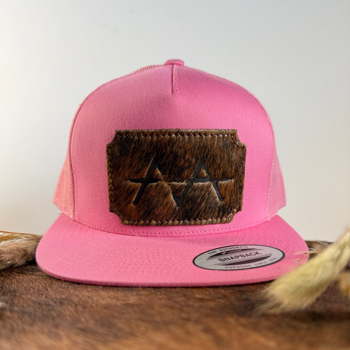 Pink trucker snapback hat with warm brindle cowhide patch and tonal brown stitched border, branded with a registered cattle brand